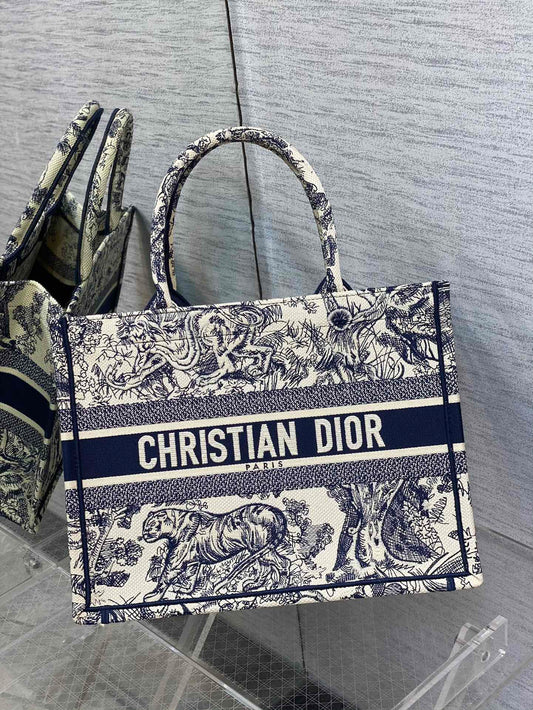 A Christian Dior Book Tote handbag in navy blue and ivory with the iconic Toile de Jouy pattern, featuring intricate wildlife illustrations and the bold "CHRISTIAN DIOR PARIS" logo across the front. Displayed on a transparent acrylic stand against a textured gray background.